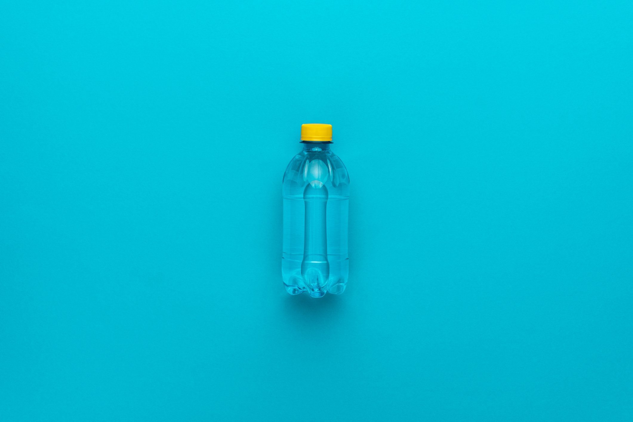 A clear plastic water bottle, standing upright, has a yellow cap against a solid turquoise background.