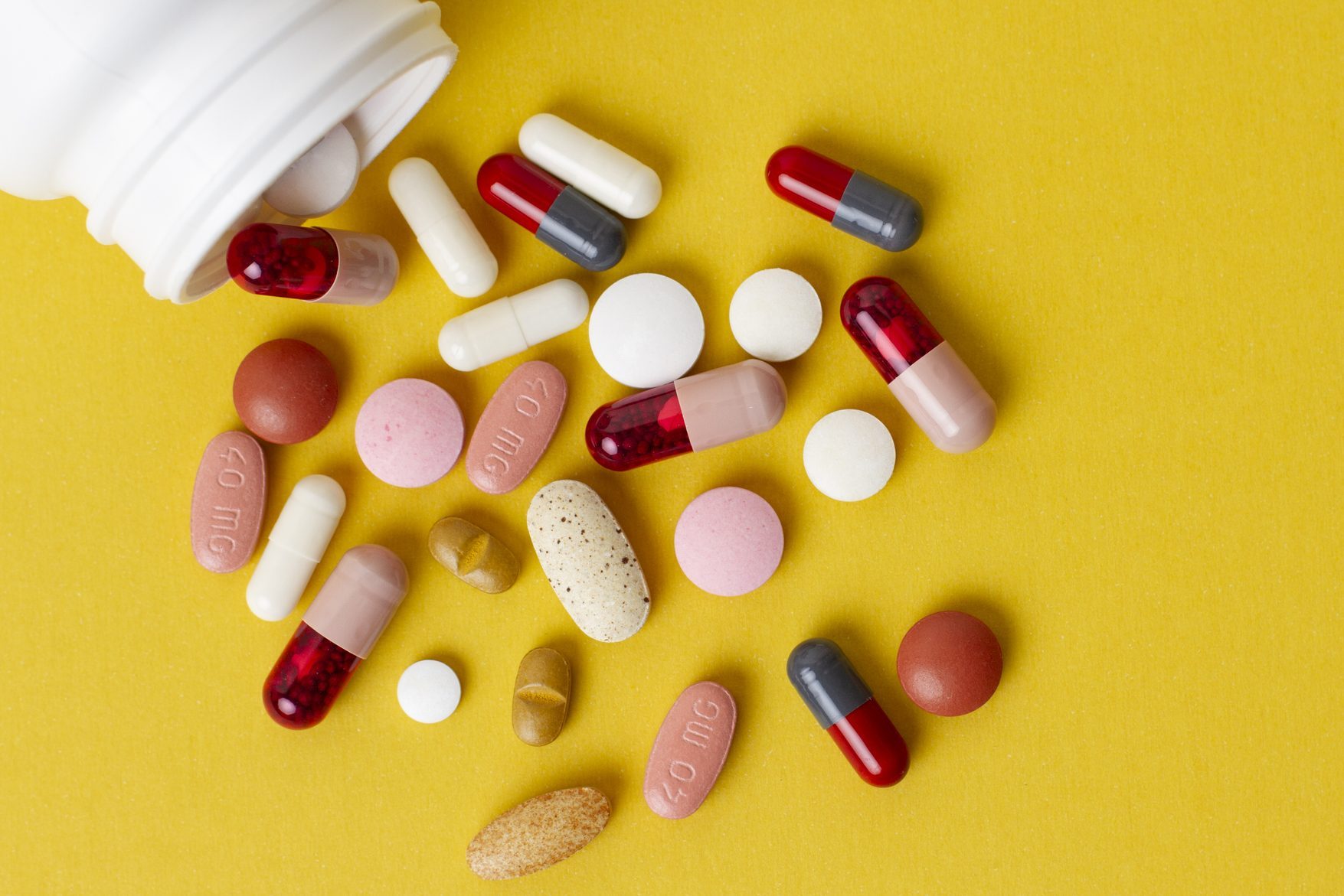 Various colorful pills and capsules spill from a white bottle onto a yellow surface, including some marked "40 mg."