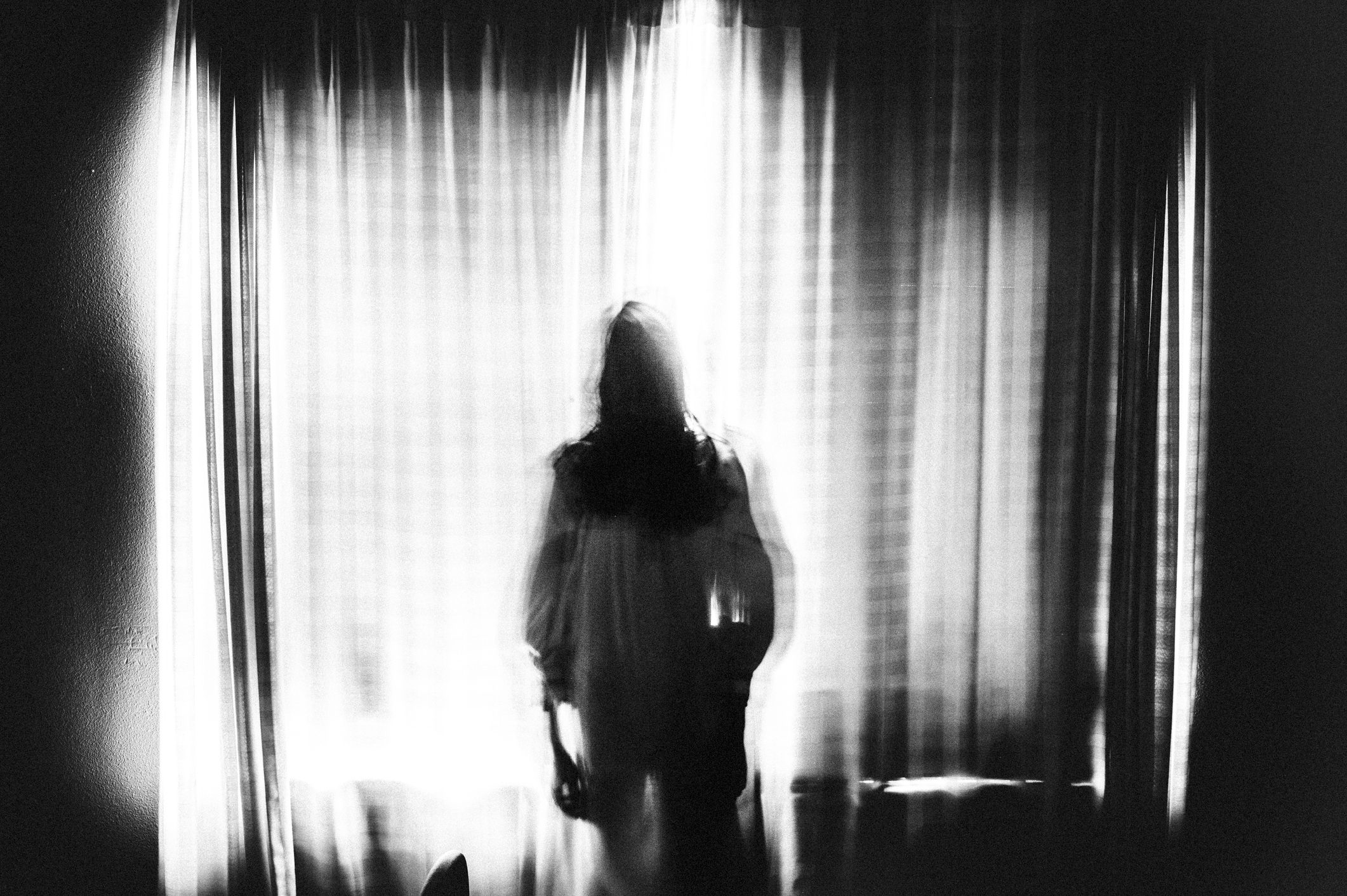 Silhouetted figure stands motionless against illuminated curtain backdrop in dimly lit room, creating a ghostly, ethereal visual effect.