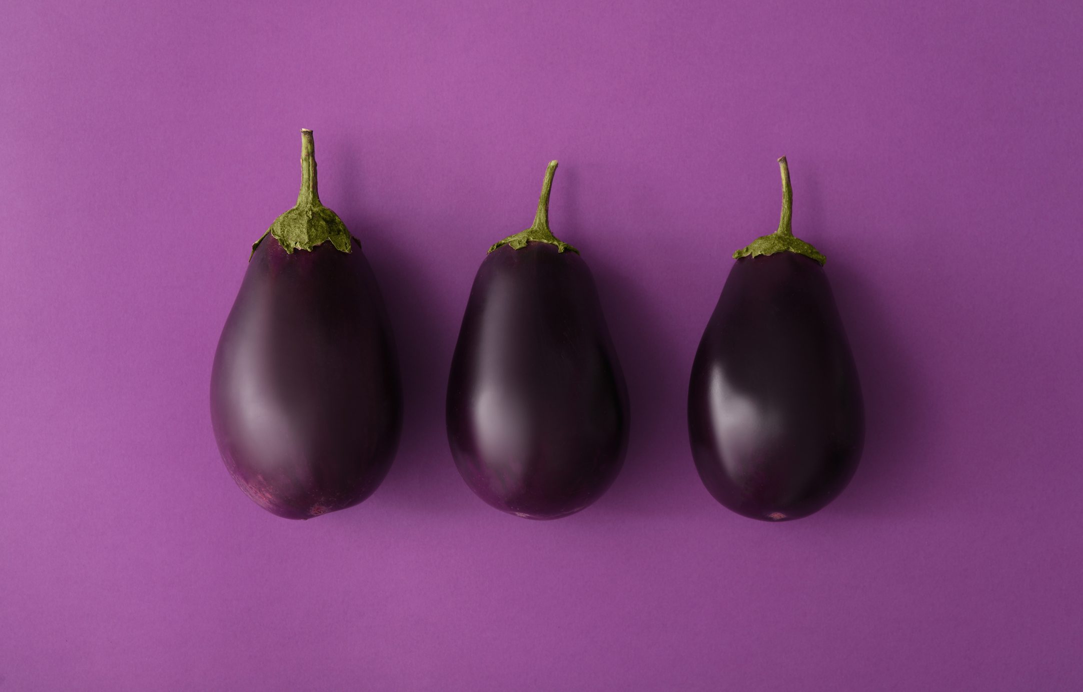 Three eggplants lie side by side on a solid purple surface.