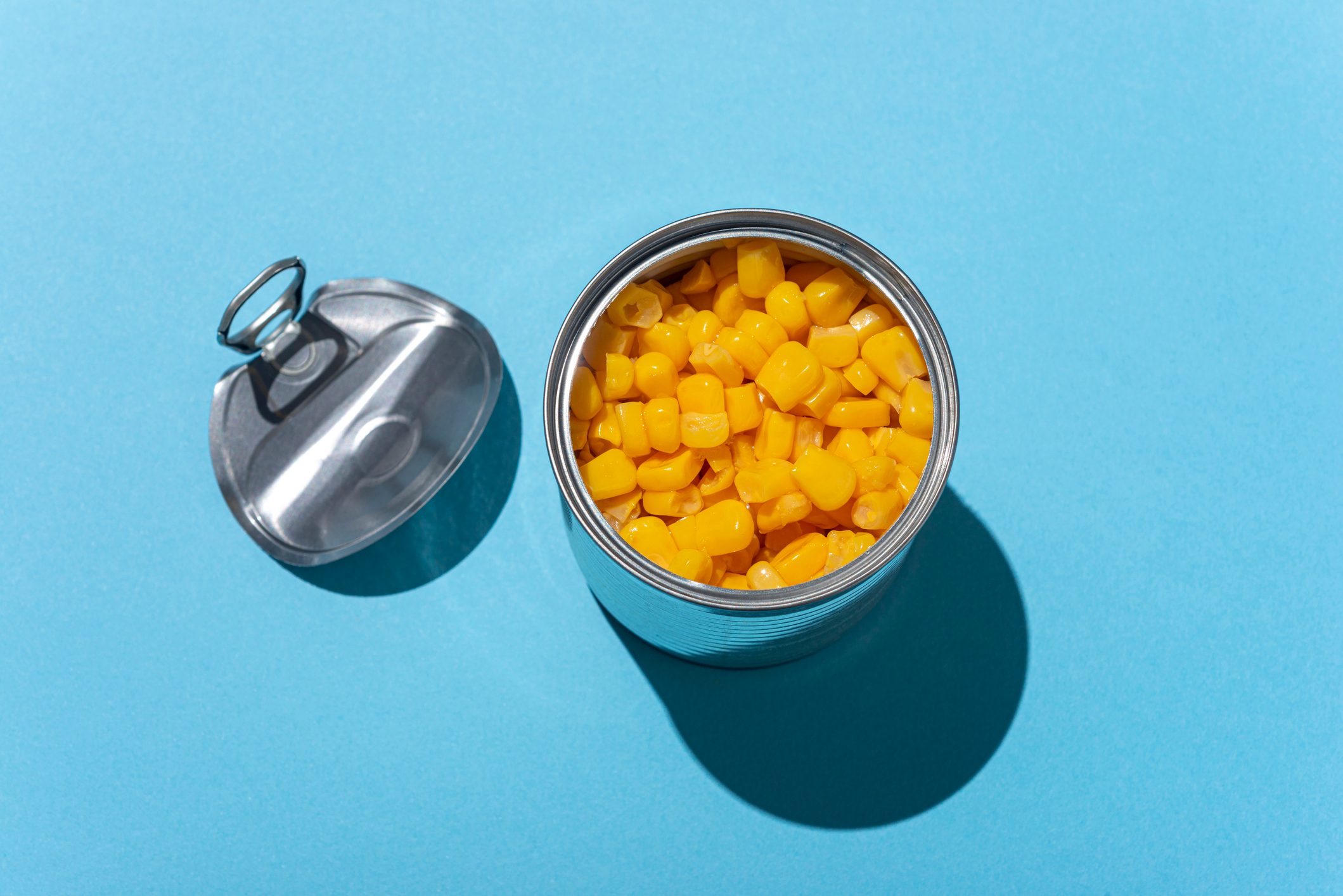 An opened can displays yellow corn kernels on a bright blue background. The detached lid lies beside it, casting a shadow.