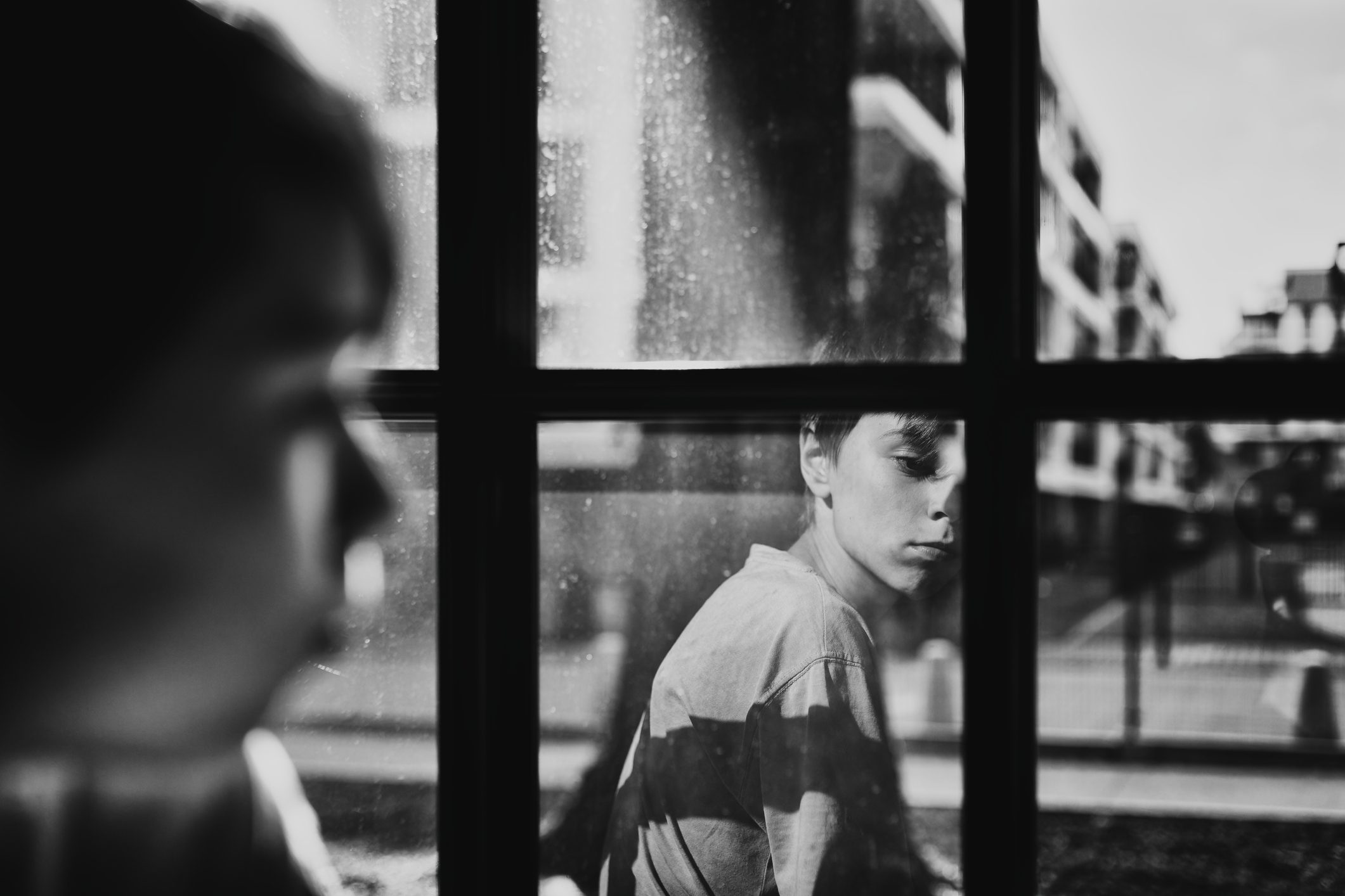 A person stares reflectively through a window, while another figure appears blurred in the foreground, set in an urban environment.