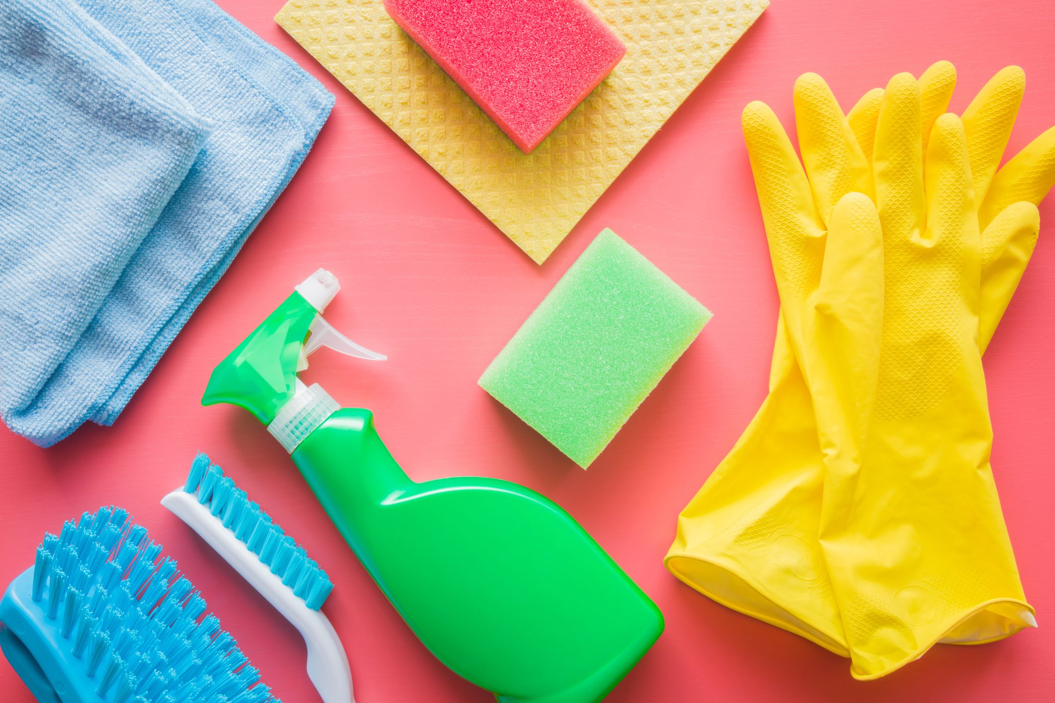 Cleaning supplies arranged neatly on a pink surface; includes yellow gloves, sponges, cloths, a spray bottle, and scrubbing brushes.