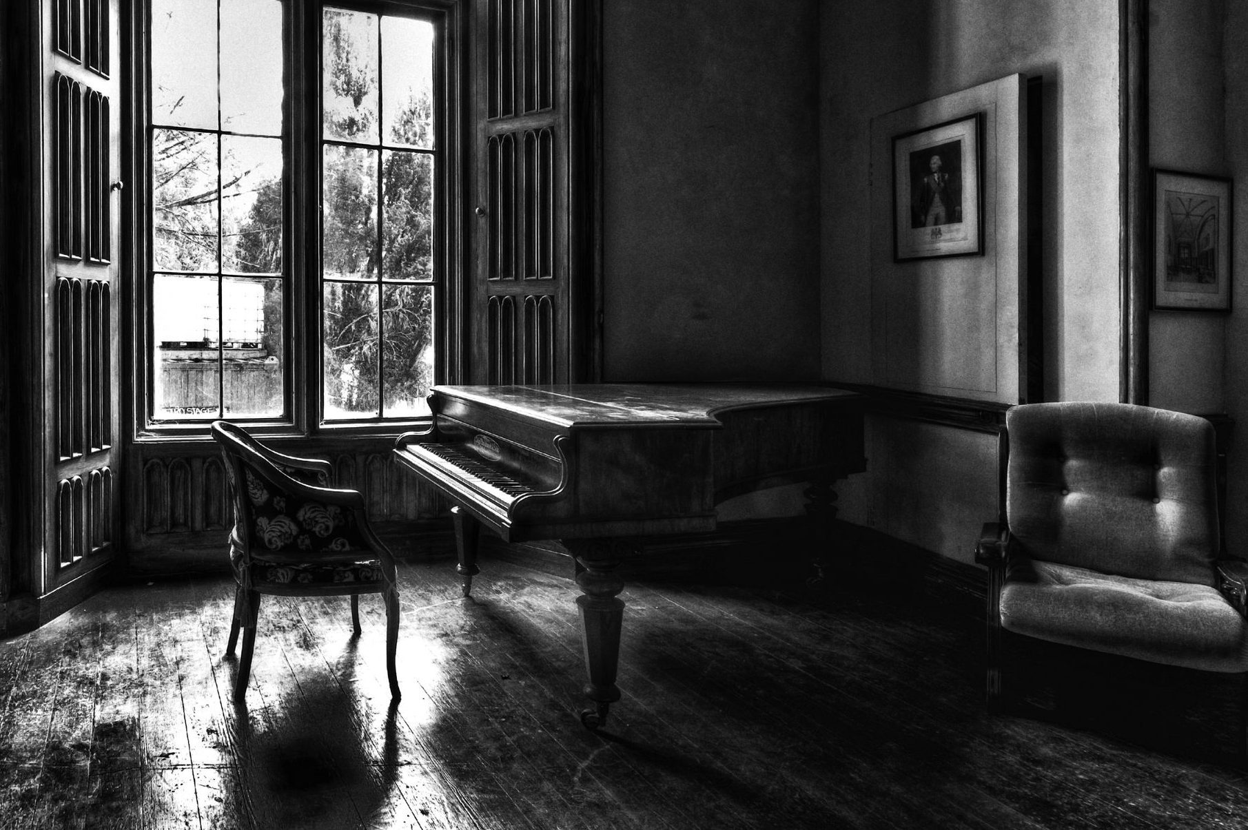 Grand piano sits unused in a dimly lit room, surrounded by a window, empty chairs, and framed pictures on the walls.
