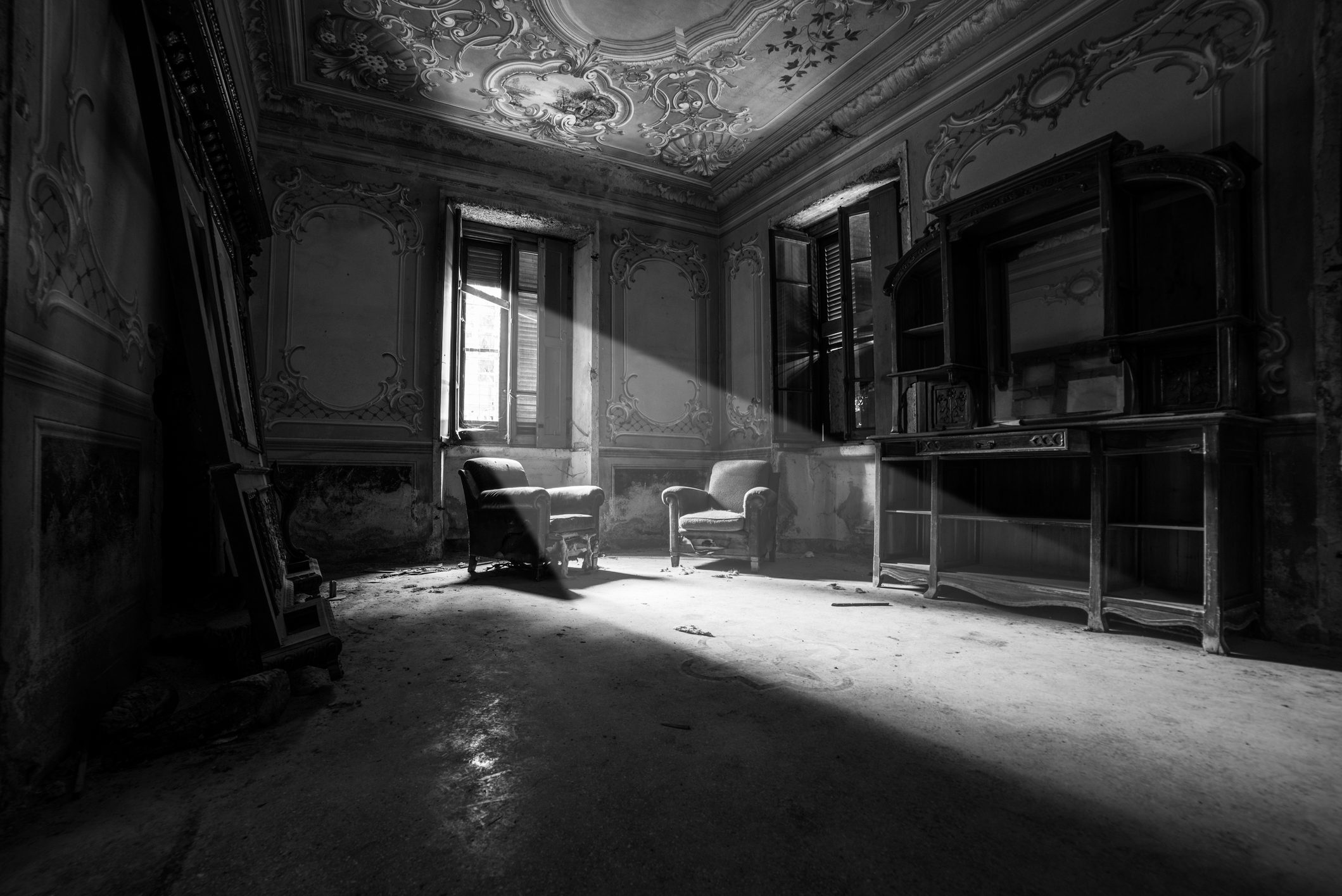 Chairs sit in shafts of sunlight within an ornate, decaying room, featuring high ceilings and sculpted walls, evoking a sense of historical abandonment.