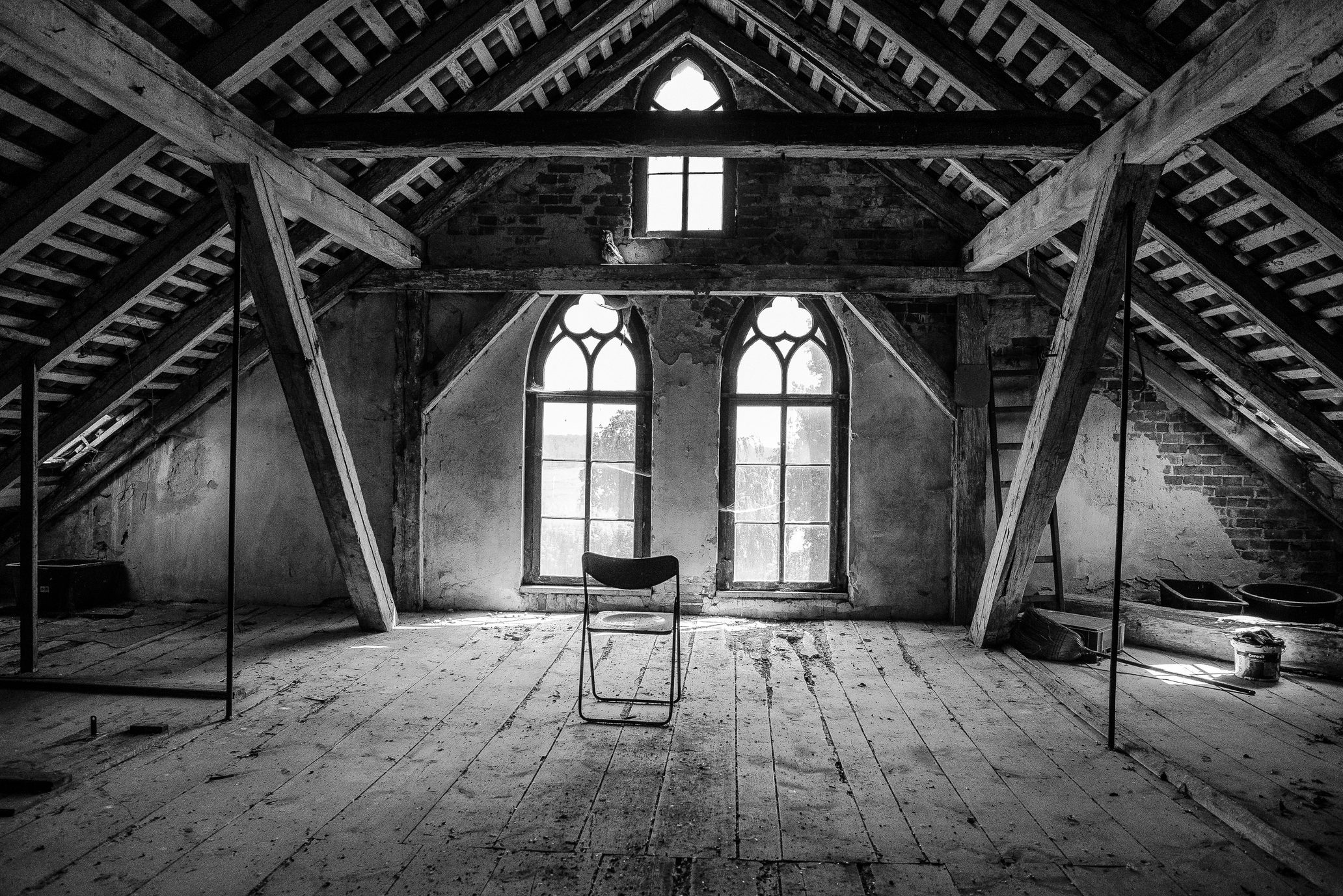 Chair sits empty, facing Gothic arched windows, in a dimly lit attic with wooden beams and plank flooring.