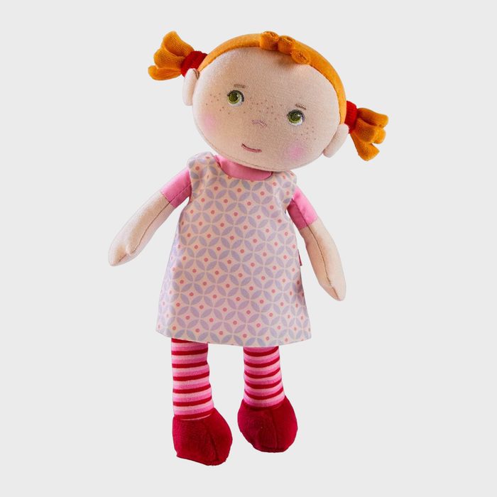 A soft doll stands upright, wearing a patterned dress and red shoes, with orange pigtails, set against a solid white background.