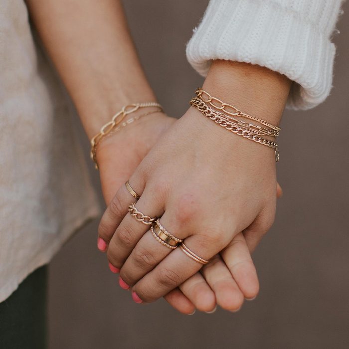 Hands clasping, adorned with gold bracelets and rings, against a neutral background.