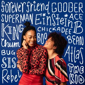 Two people smile closely in colorful clothing, set against a blue background covered with words like 