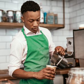 Barista pours milk into a paper cup behind a coffee machine, with shelves and jars in a tiled cafÃ© background.