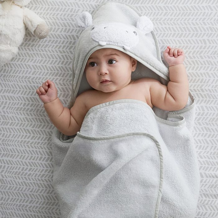 Baby wrapped in a hooded towel, lying on a patterned surface, arms slightly raised, with a plush toy nearby.