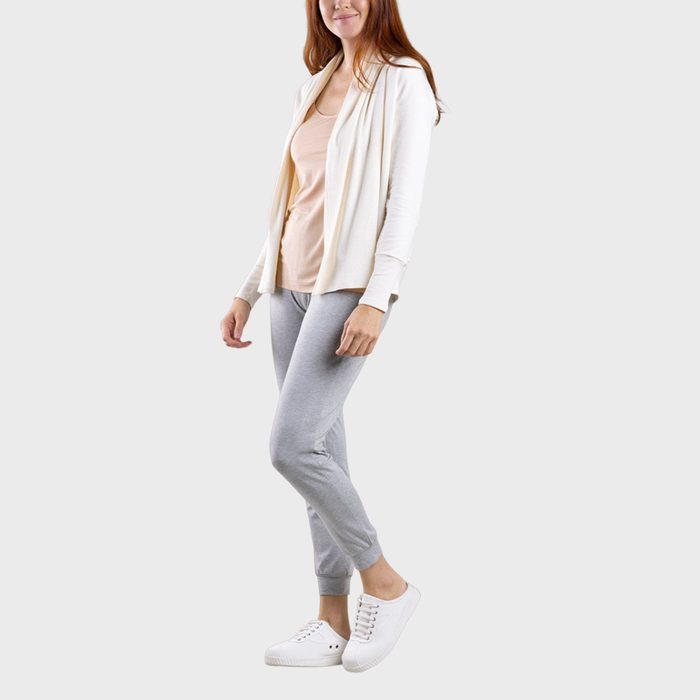 A person stands wearing a cream cardigan, light pink top, gray leggings, and white sneakers against a plain background.