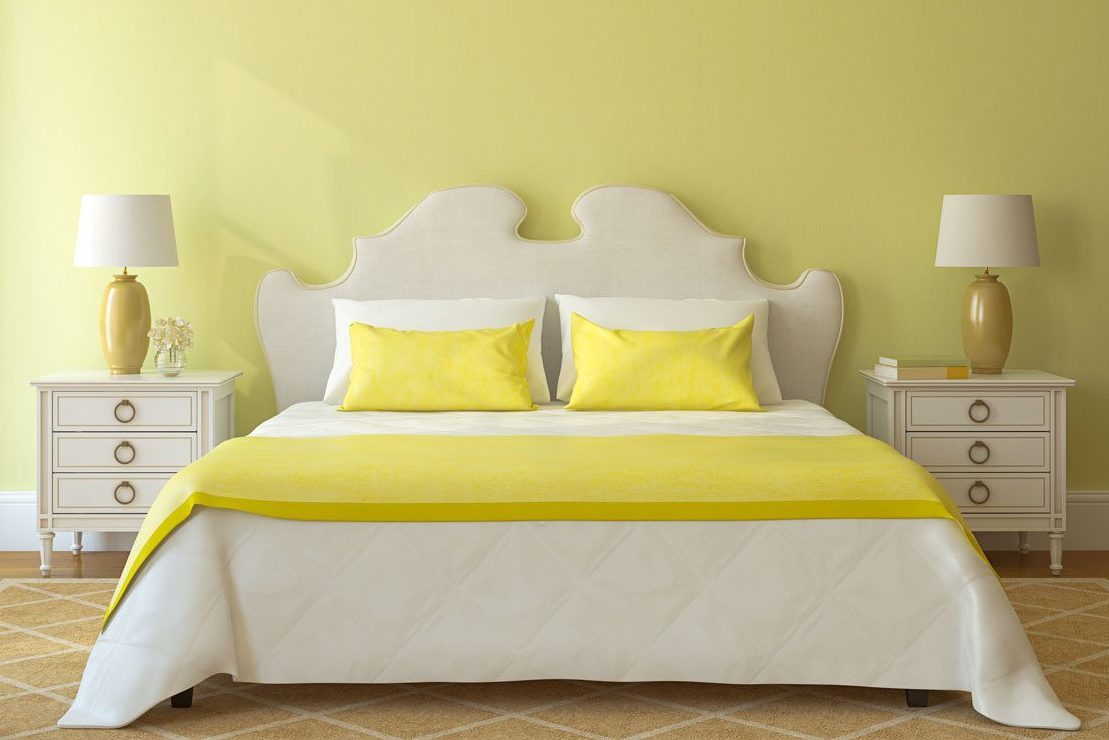 Bed with yellow pillows sits between two white nightstands, adorned with lamps, against a light green wall in a cozy bedroom.