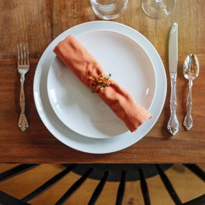 A neatly folded orange napkin rests on a white plate, placed on a wooden table, surrounded by cutlery and glassware.