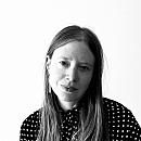 A person with long hair appears thoughtful, wearing a polka-dot shirt, set against a plain white background.