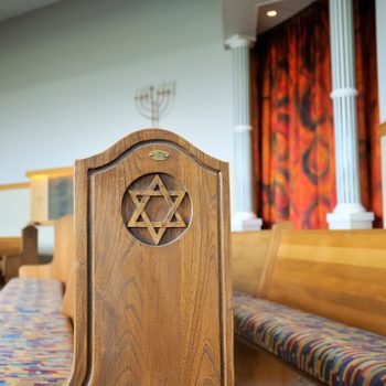 Wooden pew with Star of David carving, stands in a synagogue with colorful curtains, columns, and a menorah in the background.