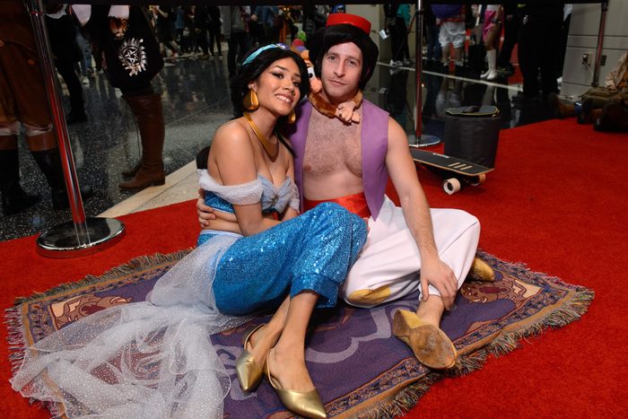 Two people in character costumes sit on a decorative rug in a crowded indoor event space.