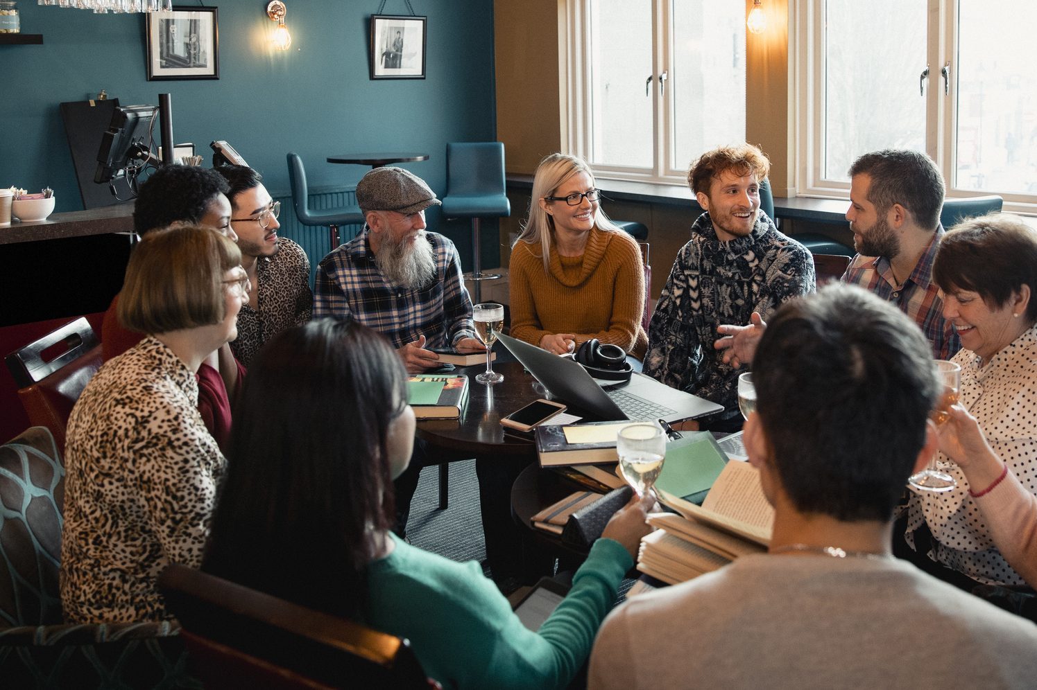 Large group of people with a mixed age range sitting together around a table. They are enjoying a drink and having a book club meeting.