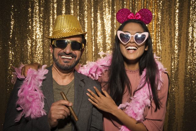 Two people wearing festive accessories, smiling and posing playfully against a shimmering gold backdrop. The scene conveys a joyful, celebratory atmosphere.