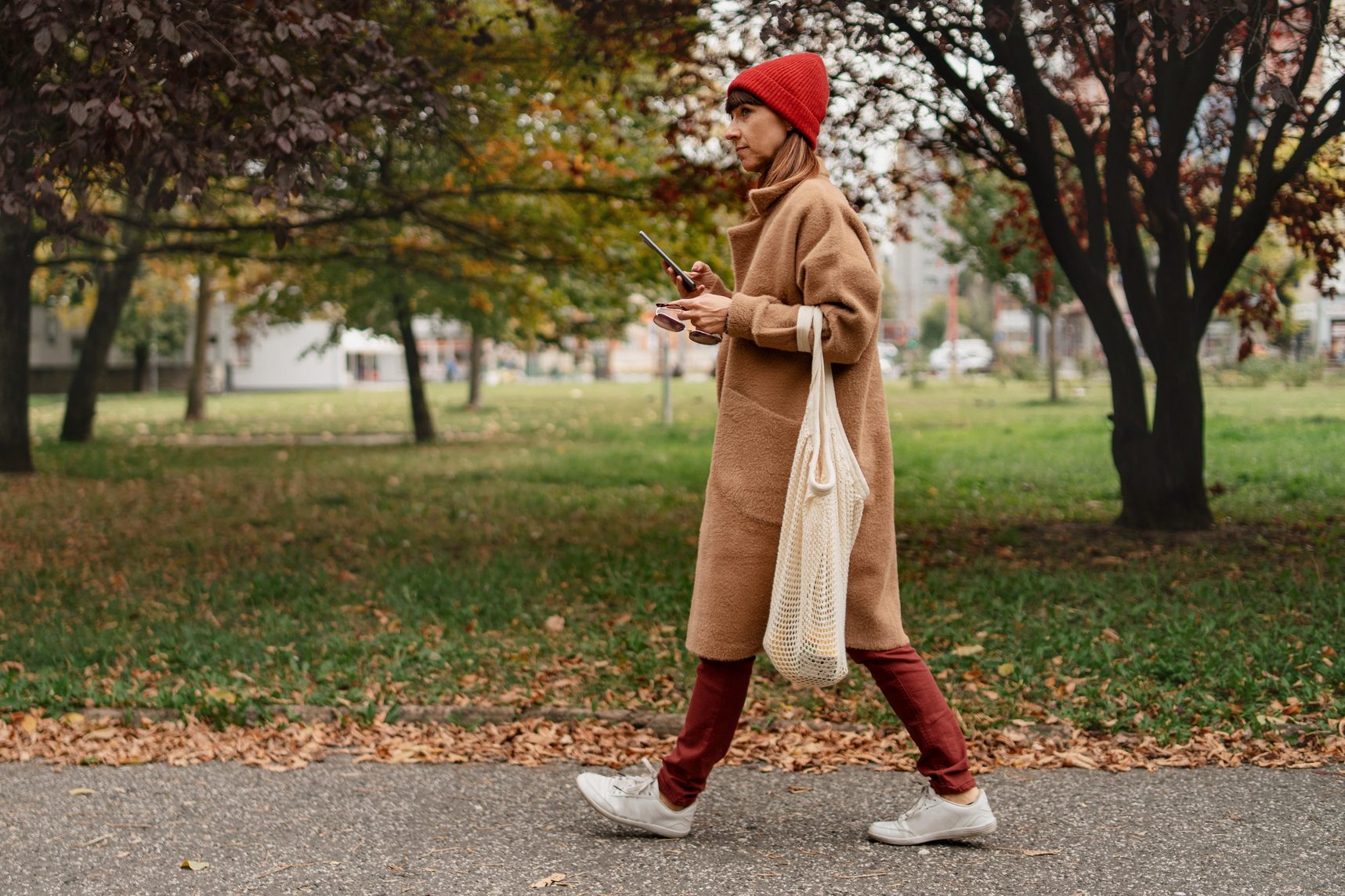 Mid adult woman with mobile phone and reusable shopping bag walking outside in the city on autumn day.