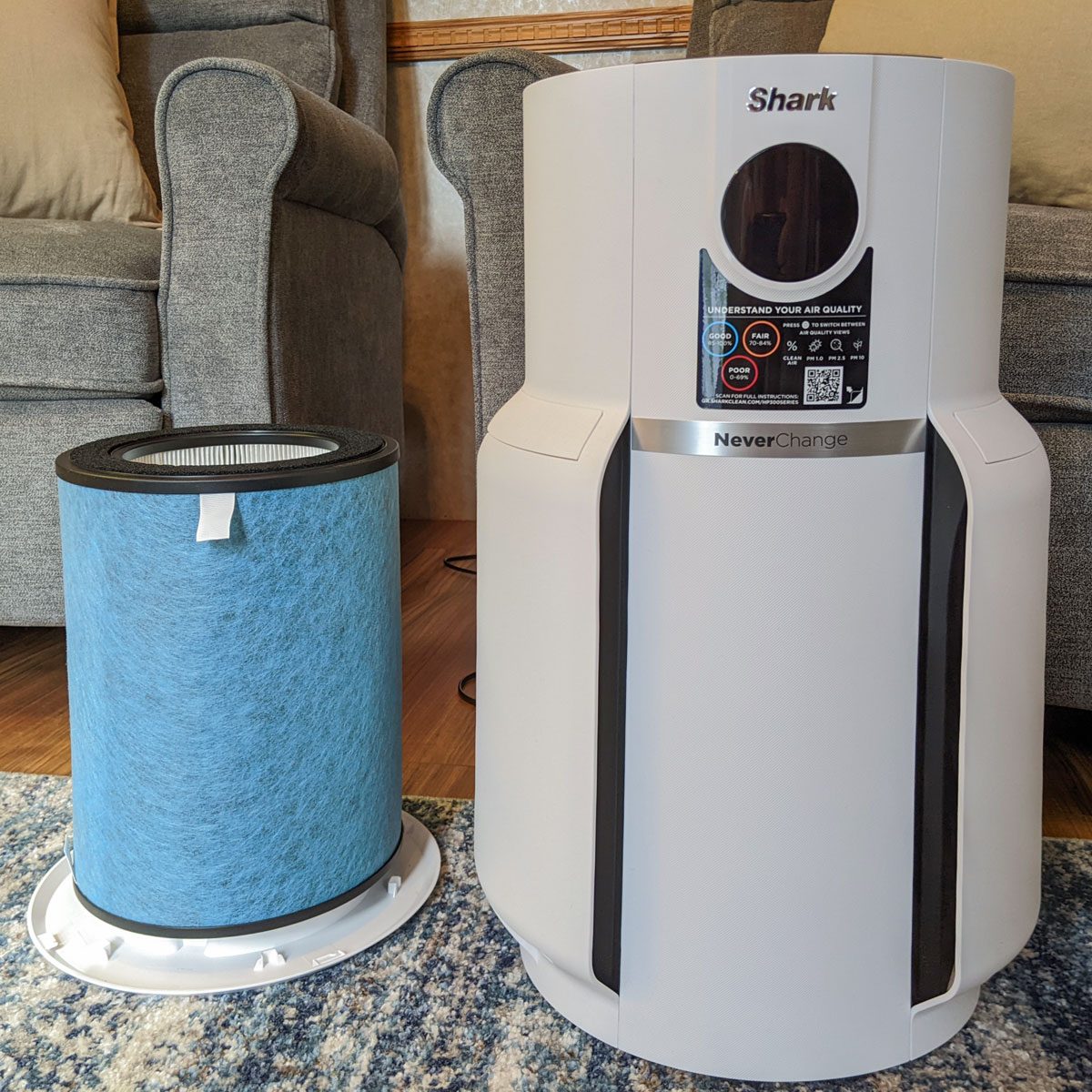 Air purifier stands next to blue filter on wooden floor, surrounded by gray upholstered furniture. Labels display air quality information.