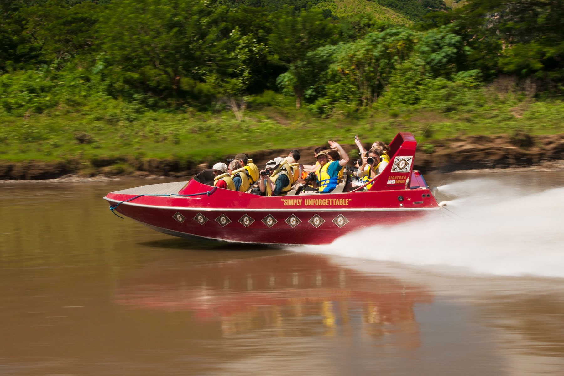 Red speedboat racing on a river, with passengers wearing life jackets, surrounded by lush green trees and vegetation. Text: "SIMPLY UNFORGETTABLE."