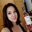 A person holds a bottle of rosé wine, smiling in front of a wooden-paneled background, with shelves of bottles visible behind.