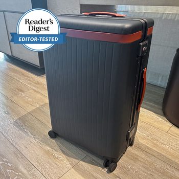Suitcase stands upright on wooden floor; black with orange accents, caster wheels, and visible lock. "Readerâ€™s Digest Editor-Tested" badge in upper left corner.
