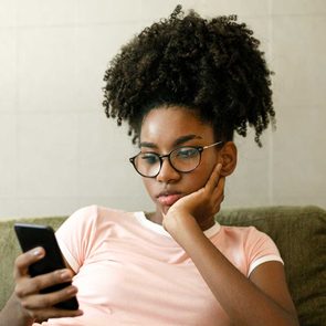 Person holding a smartphone, sitting on a couch, looking intently at the screen with a thoughtful expression in a simple indoor setting.