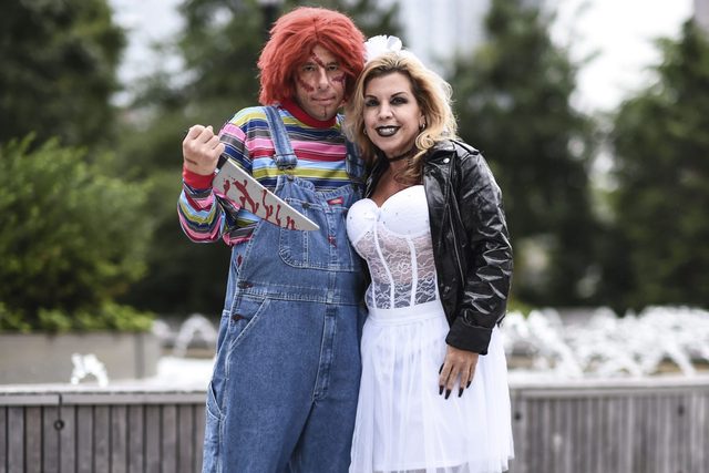 Two people in costume pose; one has red hair holding a knife, the other in a lace bodice and leather jacket. Outdoor, greenery background.