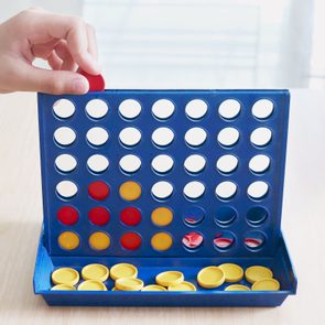 Hand places red disc into a blue Connect Four board, surrounded by yellow and red discs on a light wooden table.