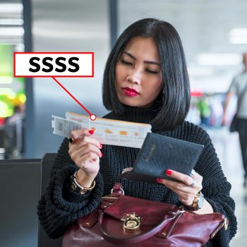 A woman examines a boarding pass marked with "SSSS" at an airport terminal, seated with luggage and other travelers around her.