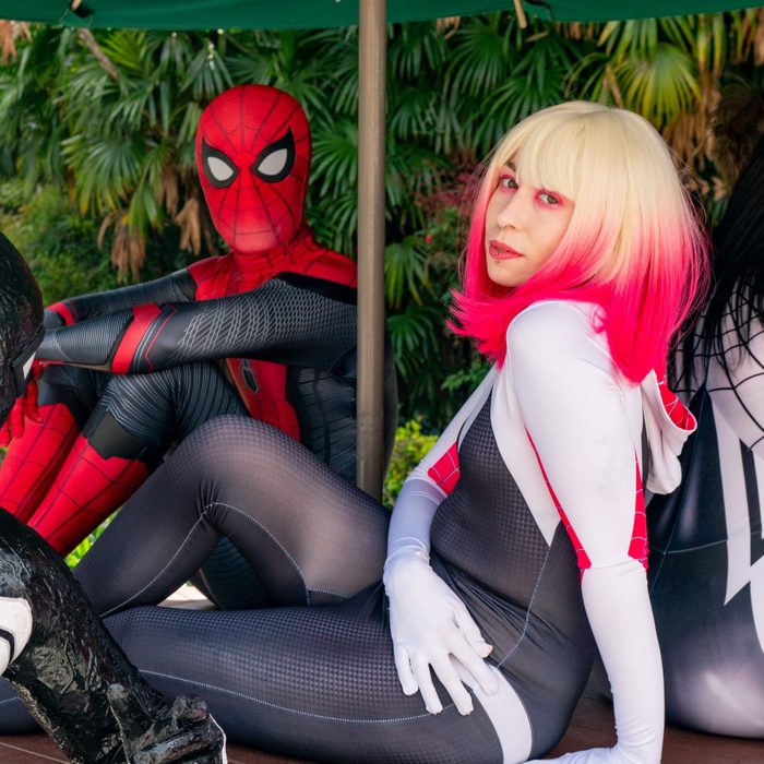 Cosplayers sit relaxed, dressed as Spider-Man and Spider-Woman, under a green umbrella with lush greenery in the background.