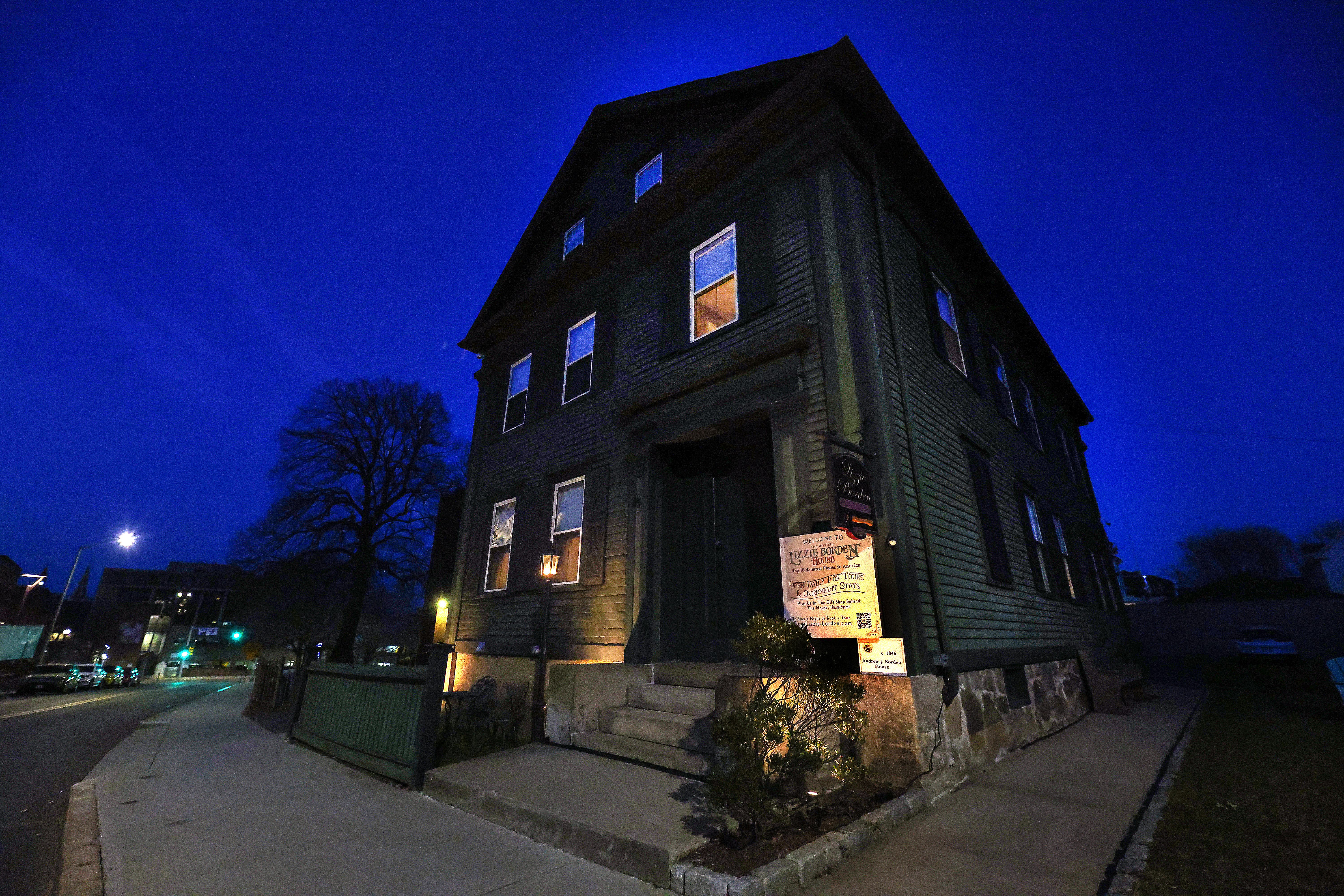A large, dark wooden house with lit windows stands on a dimly lit street corner at night, under a clear, deep blue sky.