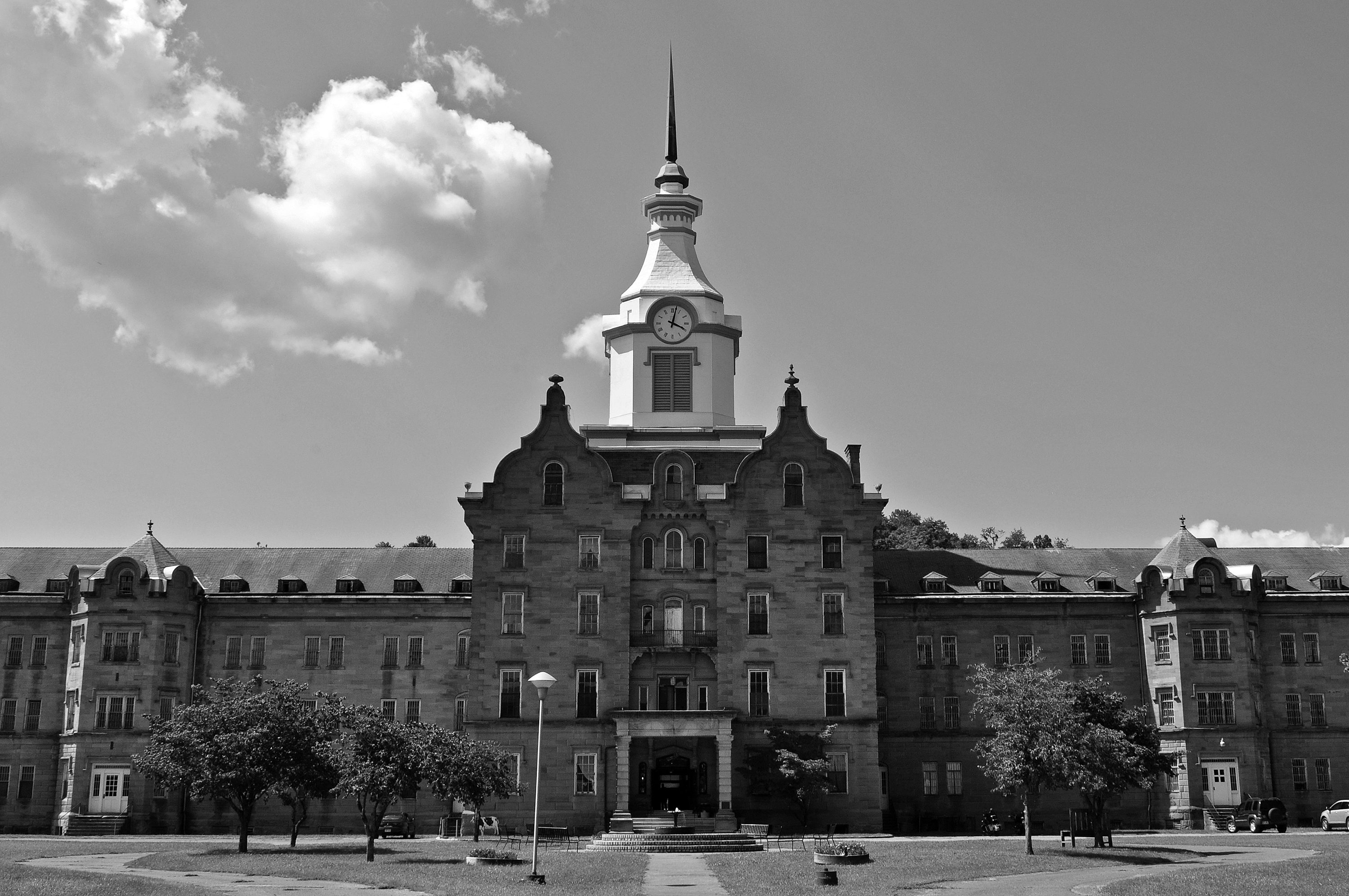 A large building stands under a cloudy sky, featuring a clock tower and surrounded by a few trees on a grassy area.