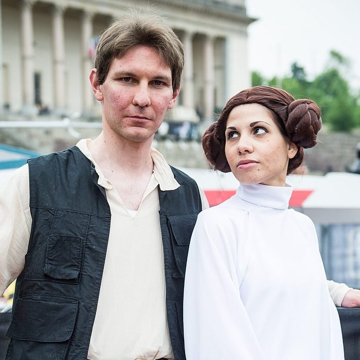 Two people pose in sci-fi costumes, one in a black vest and the other in a white gown, outside a classical building.