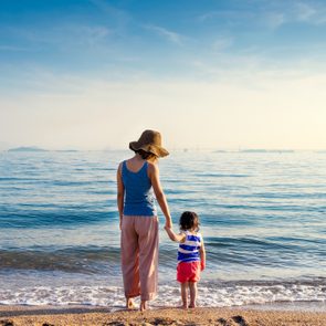 Two people stand hand-in-hand by the sea, facing gentle waves under a clear blue sky.