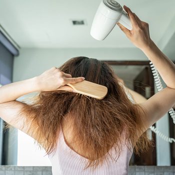 Person brushes hair while using a hair dryer; standing in a bathroom, wearing a sleeveless top.