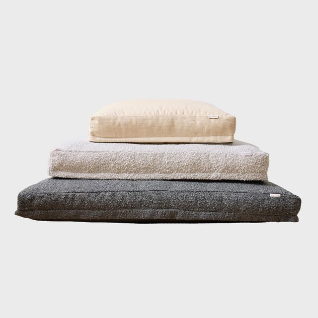 Three stacked cushions of varying sizes. The top is beige, middle is light gray, and bottom is dark gray. Simple background.