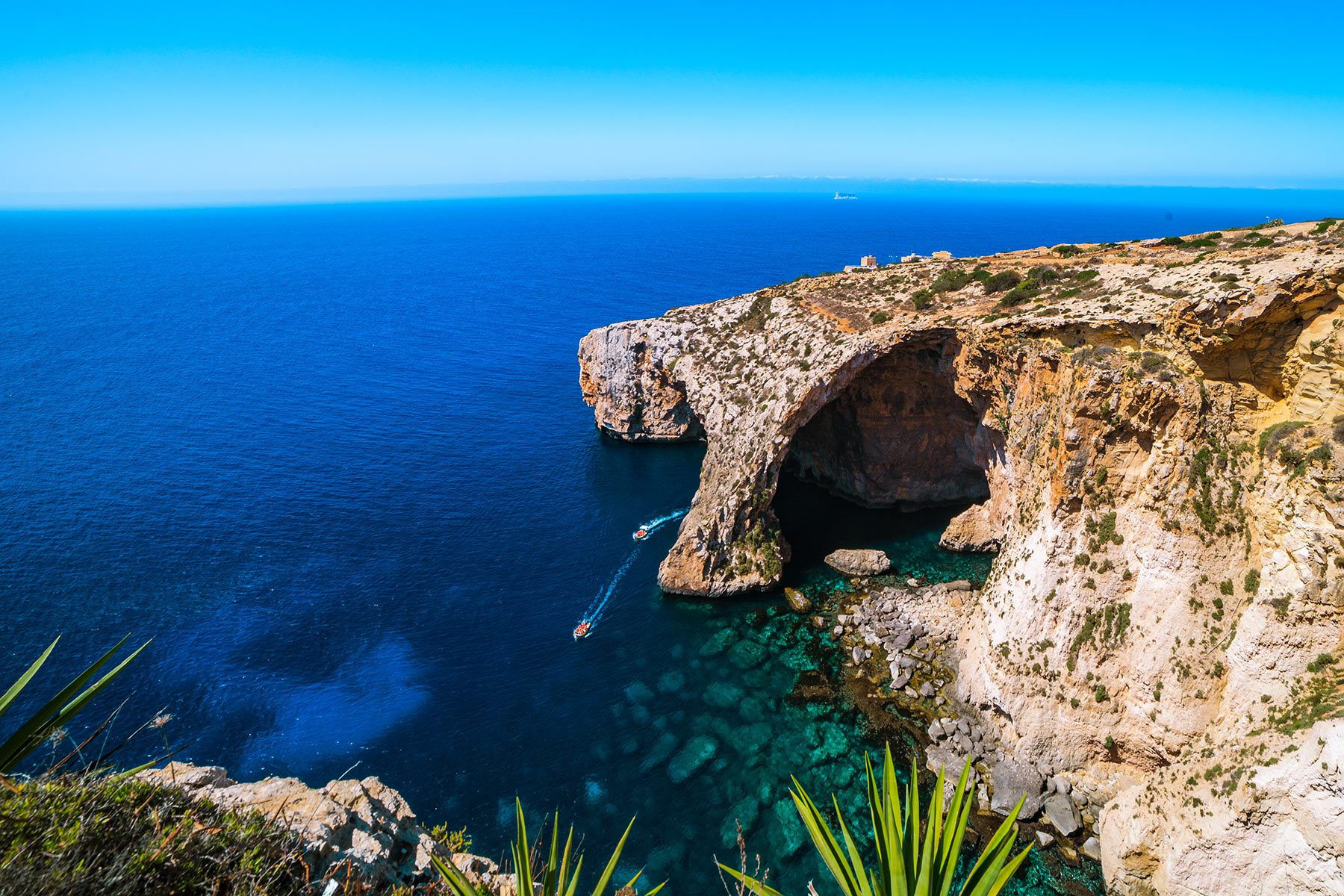 Boat navigates azure waters beside rocky arch cliffs under a clear blue sky. Sparse coastal vegetation surrounds the scene.