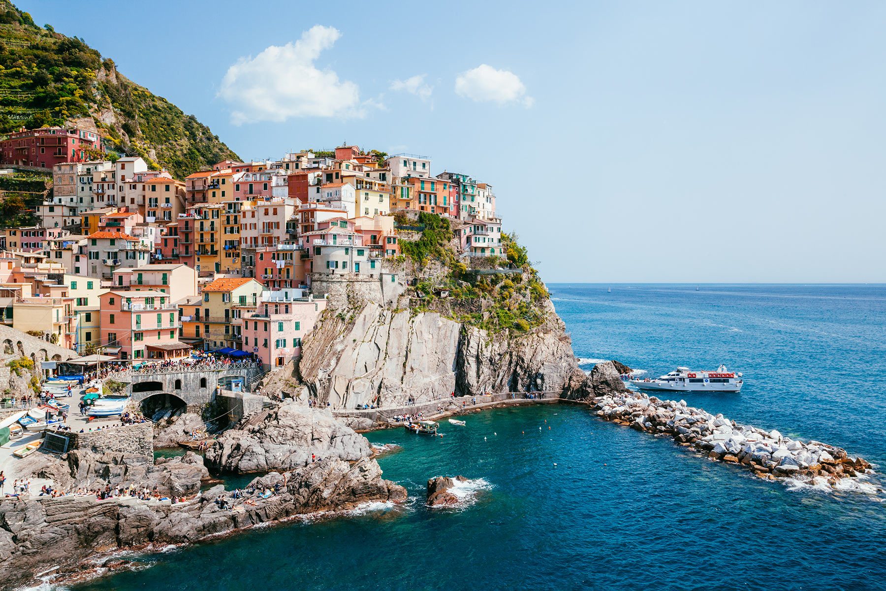 Colorful buildings perch atop a rocky cliff overlooking a calm blue sea, with boats docked nearby under a clear sky.