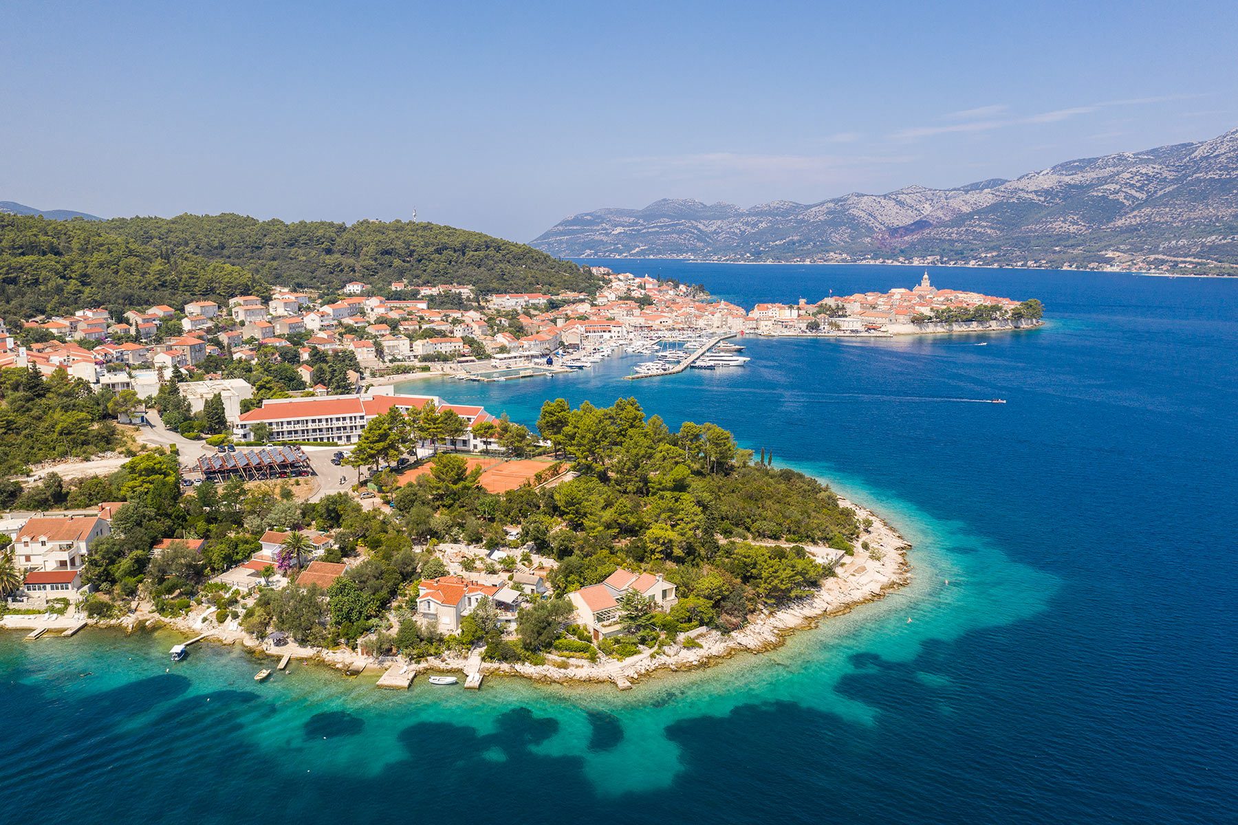 A peninsula with red-roofed buildings is surrounded by turquoise water, adjacent to a marina, with distant mountains under a clear sky.
