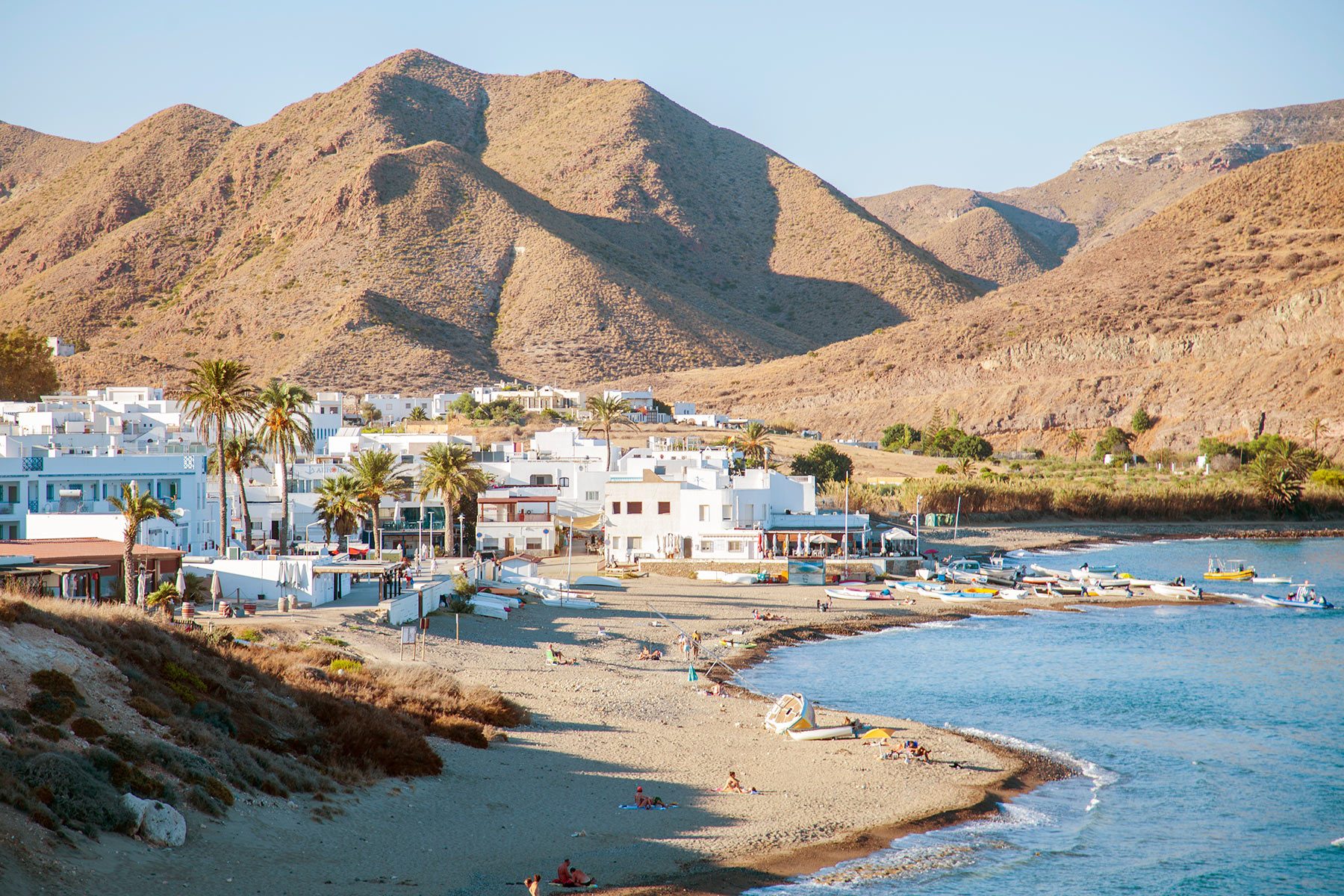 Houses rest nestled against rocky hills. People relax on a sandy beach beside the calm sea. Sunlight illuminates a peaceful coastal village.