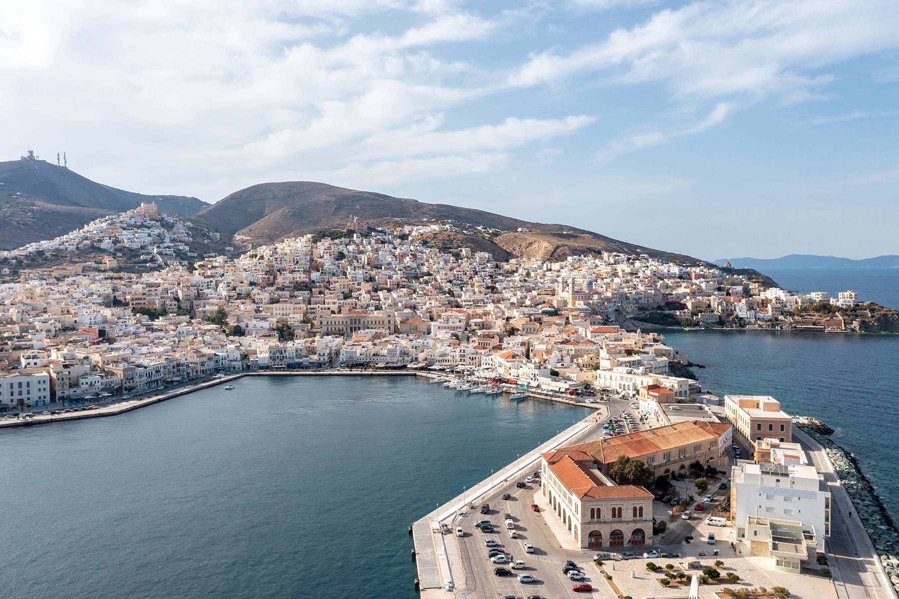Buildings cluster on a hillside, overlooking a calm bay, surrounded by mountainous terrain under a cloudy sky.