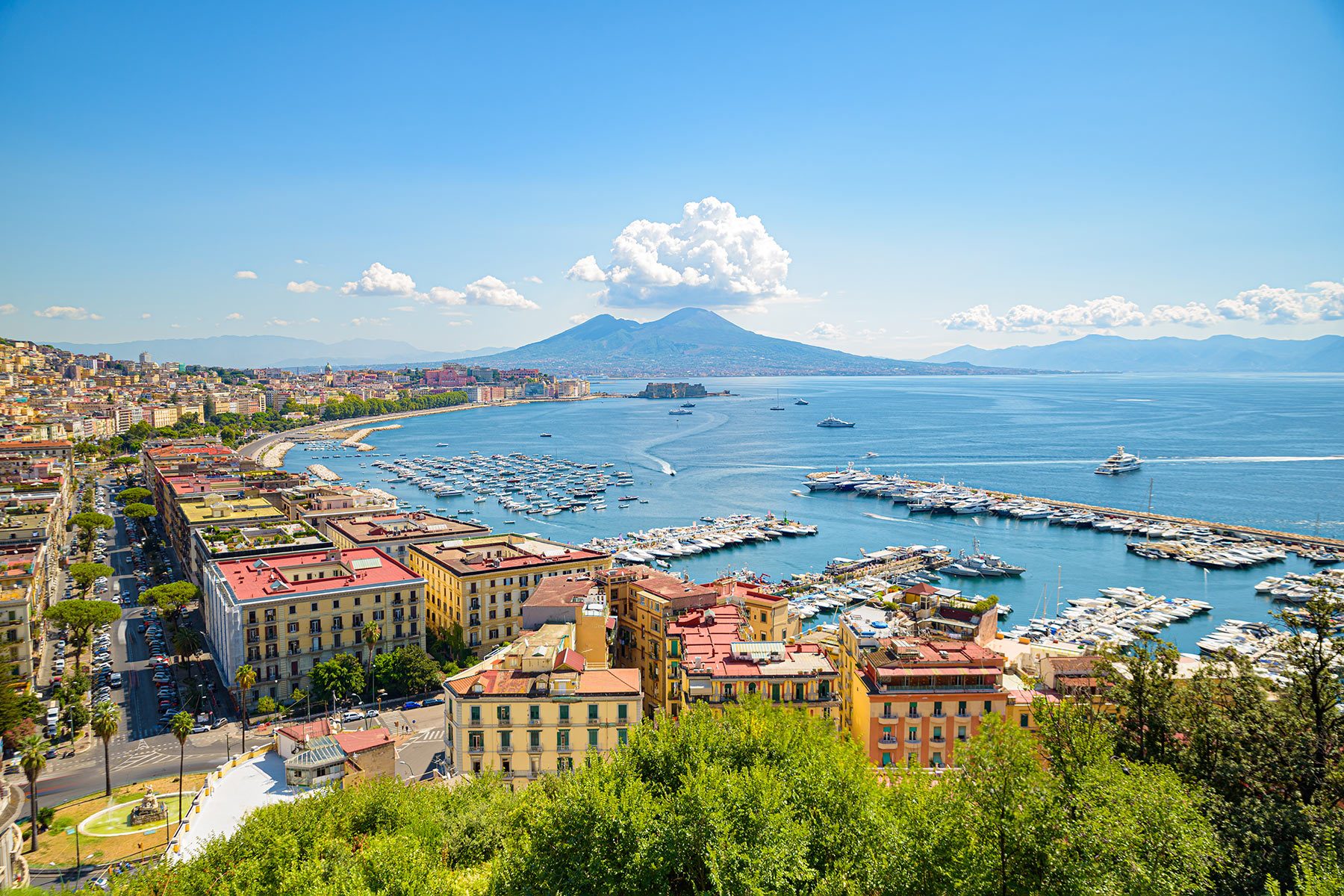 Cityscape overlooks vibrant harbor with boats, bordered by colorful buildings and lush greenery, under clear blue sky with distant mountain view.