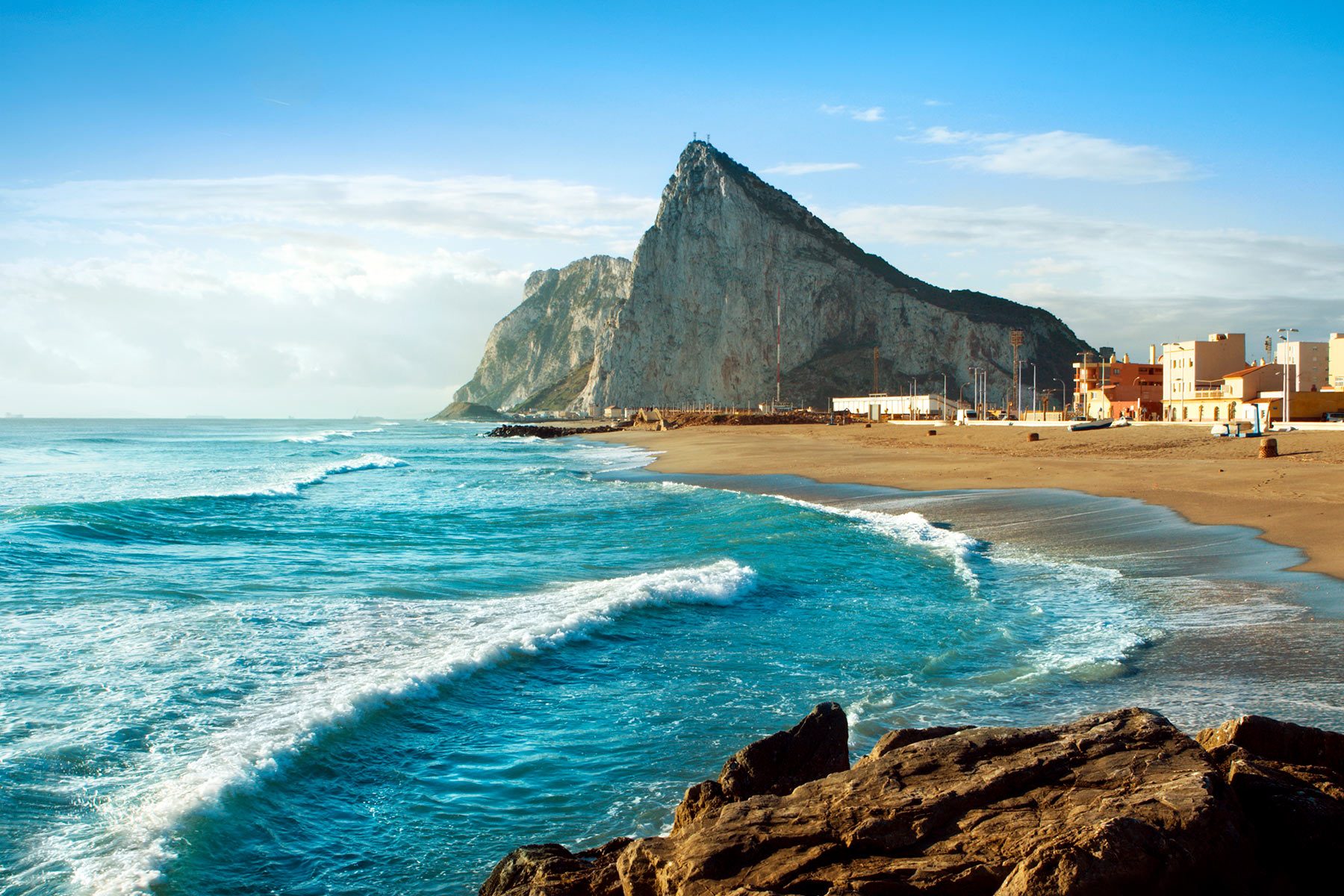 Rocky mountain dominates the seascape with waves crashing on the sandy shore, buildings in the distance under a clear blue sky.
