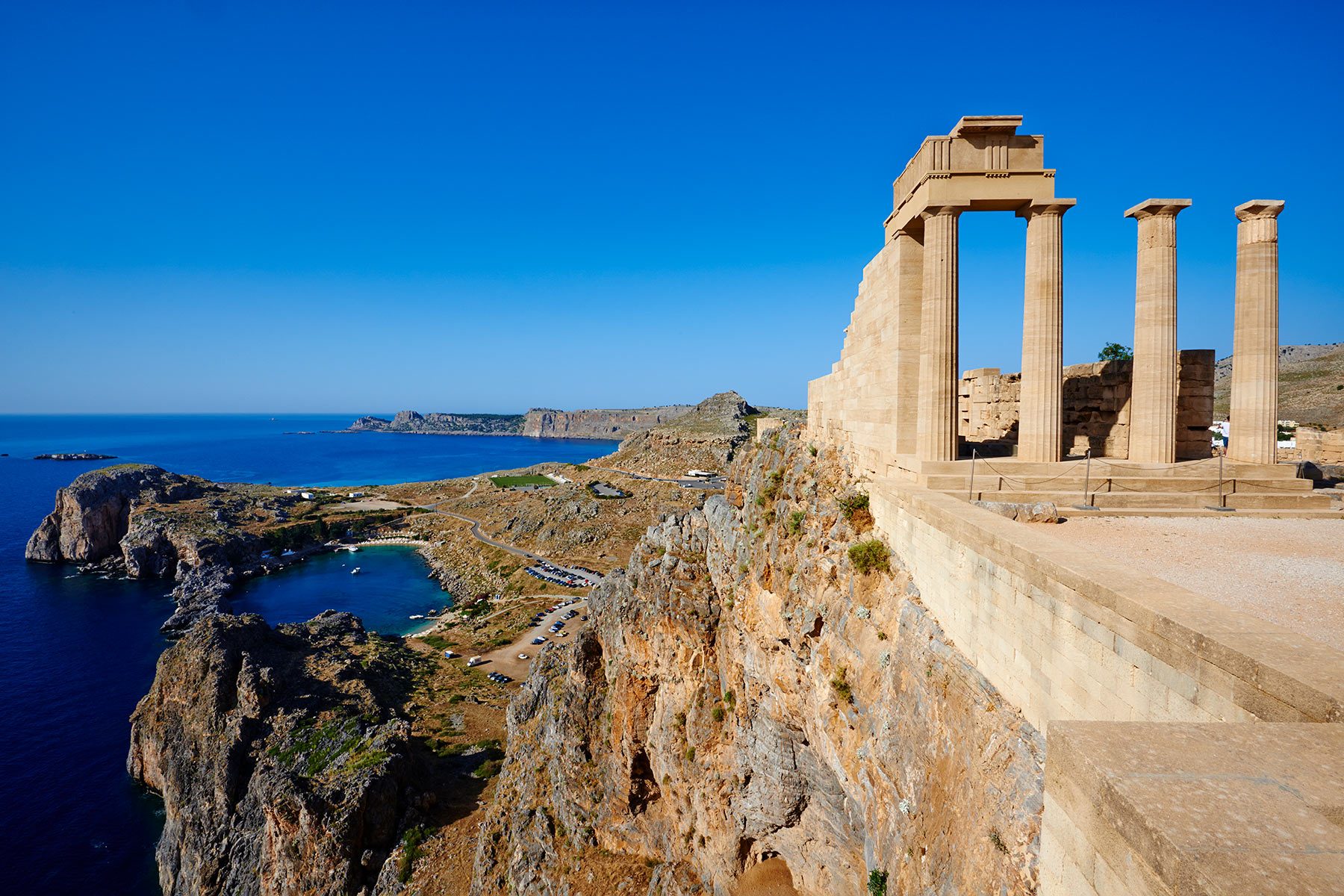 Ancient stone columns stand atop a rugged cliff overlooking a vast blue sea and coastline under a clear sky.