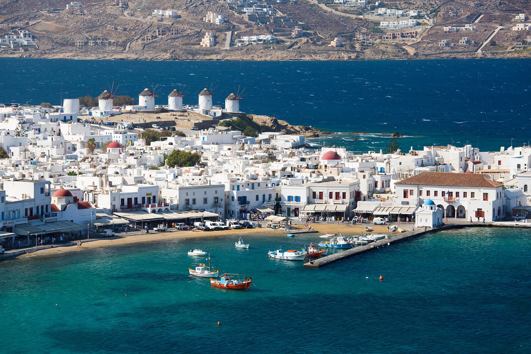 White buildings rest by the turquoise sea, with several windmills on a hill, under a clear blue sky.