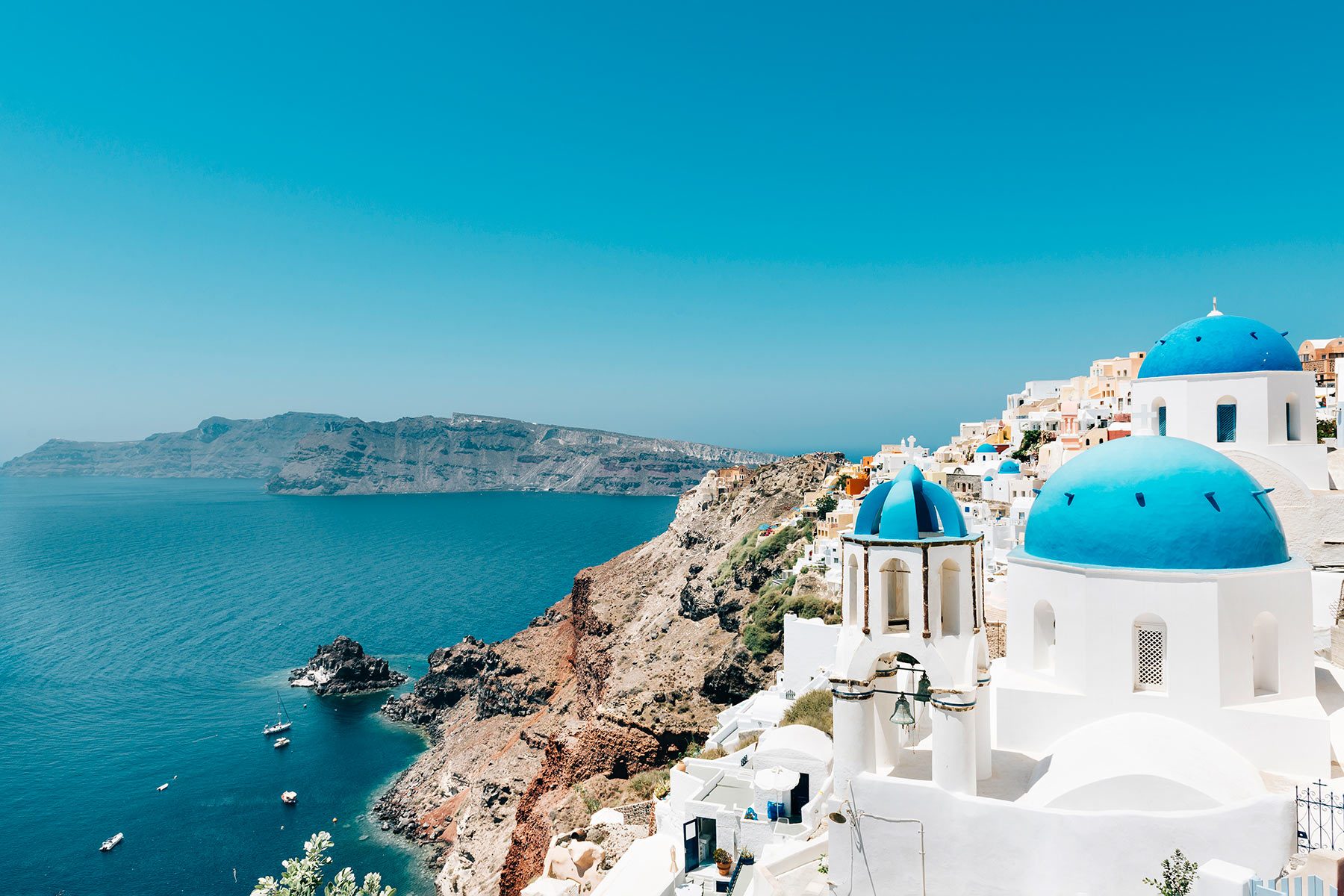 White buildings with blue domes on a cliff overlook the sea, under a clear blue sky, with distant hills in the background.