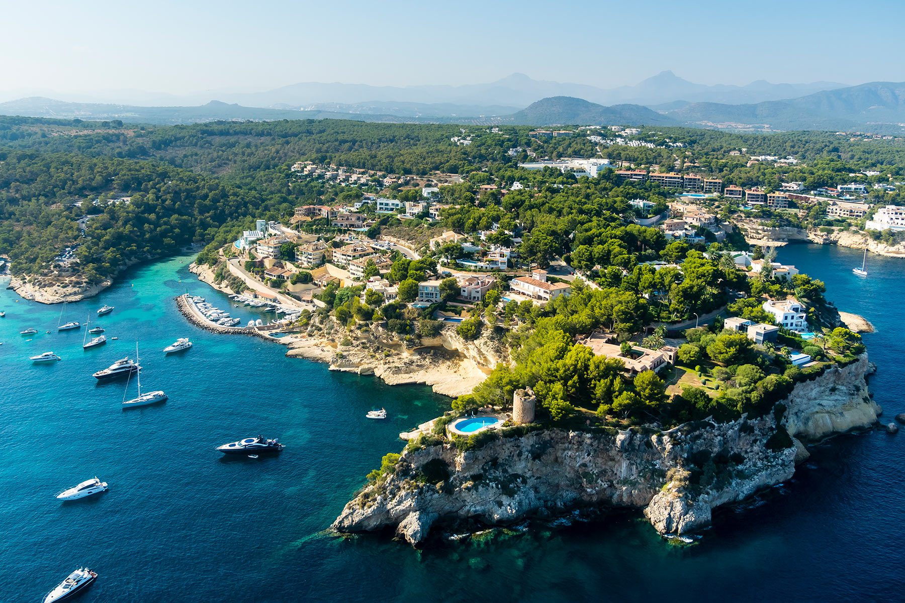 Boats float in a blue sea near a lush, hilly peninsula with houses and trees, set against distant mountains under a clear sky.